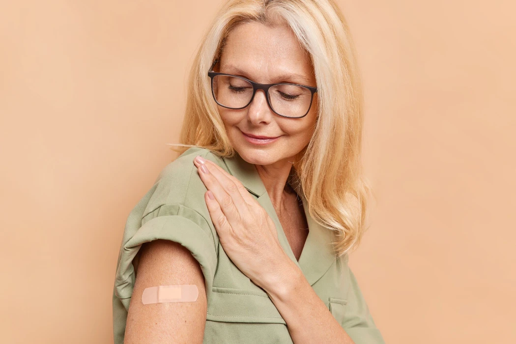 Sideways shot of adult woman with fair hair looks attentively at arm with plaster after vaccine injection protects herself from coronavirus wears spectacles and dress isolated over beige wall